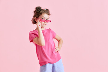 Portrait of beautiful little girl child in casual clothes, sunglasses posing against pink studio background. Concept of emotions, childhood, education, fashion, lifestyle, ad