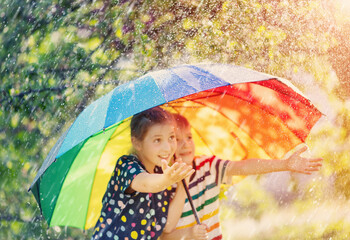 Boy and girl standing outdoors in rainy day under colourful umbrella and trying to catch any...