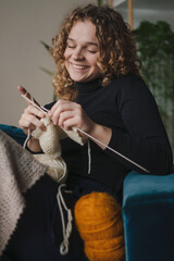 Positive young female with curly hair smiling and looking at camera while sitting on sofa and weaving a scarf