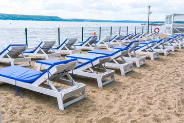 Wooden chaise longue for relaxing on the beach with a soft removable mattress. Relaxing on the beach by the river.  Summer tan by the water. A relaxation area with sun loungers on the sandy beach.