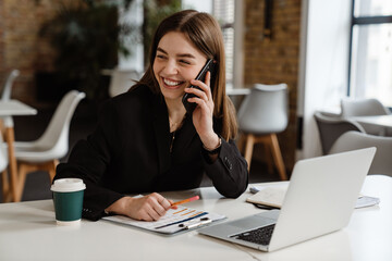 Cheerful business woman talking on mobile phone while working on laptop in office