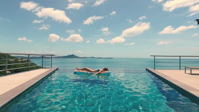 Outdoor lifestyle portrait of young pretty sexy girl swimming with neon bright swimming mattress at the pool, wearing bikini and sunglasses, view on ocean, relax and having fun on vacation.