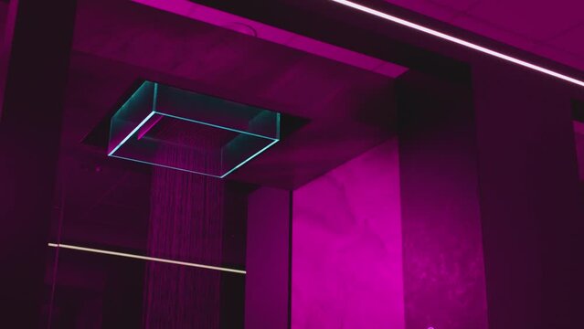 Water flowing from the beautiful modern designed shower . Overhead  futuristic , luxury shower in dark bathroom . Pink , purple led lights inside . Unique design . Water falling , dropping , pouring