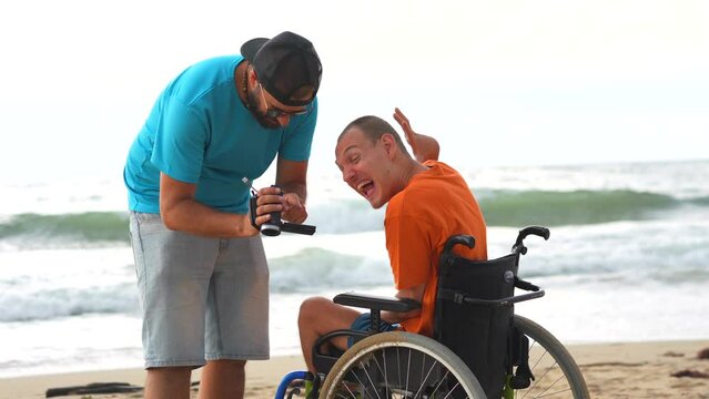 A disabled person in a wheelchair on the beach having fun with a friend and a video camera, taking a selfie