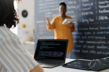 Rear view of student typing codes on computer while sitting at desk at lesson with teacher explaining material in background