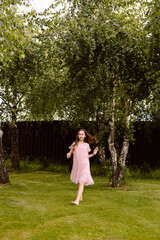 A little girl with long brown hair in a long pink dress dances and jumps on a well-trimmed lawn in the garden of a small house in the village in summer