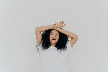 Fototapeta premium Overjoyed curly-haired lady, hands on forehead, laughing with mouth wide open, energetic vibes. Casual attire, white studio backdrop. Hilarious joke!