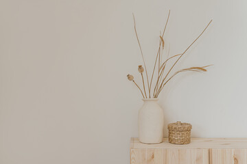 Aesthetic minimal home interior design. Clay jug with dried grass and poppy stems, rattan casket on wooden table over white wall. Luxury bohemian home interior decoration