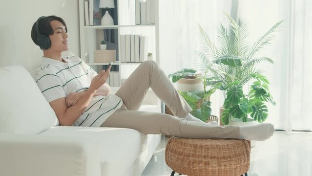 Youth attractive Asia male with casual cloth calm feeling lay down on sofa wear headphone hold smartphone listen music playlist relax at living room in cozy house. Lifestyle leisure at home concept.