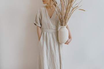 Woman in sandy beige dress holds earthenware with dried pampas grass over white wall. Aesthetic...