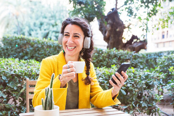 Smiling woman with her phone holds a cup of coffee in a park with headphones on