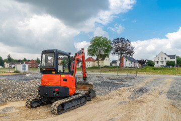 Rear view of a small red excavator standing on the edge of a construction site. Blue sky with white...