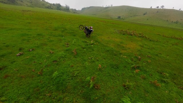 Enduro motorbike biker speed riding extreme stunt tricks up front wheel on green valley field aerial view. FPV sport drone close shot male racer standing on back wheely motorcycle adrenalin travel 4k