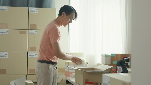 Young attractive Asia male entrepreneur E-commerce small business prepare product put in paper box hands hold packing machine and sealing cardboard boxes with duct tape at small online retail shop.