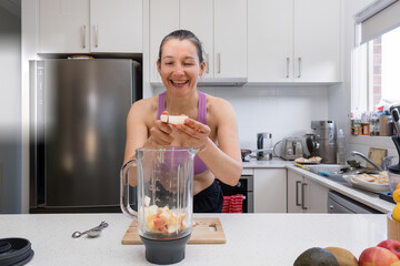 Smiling woman in sport bra adding fruit to a blender to make smoothie.