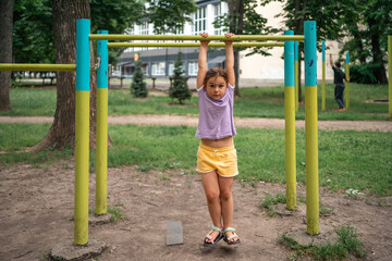 Fototapeta na wymiar 5 years old girl doing sports on the outdoors workout zone. Pull-ups on the bar, stretching back and spine, healthy lifestyle for kids