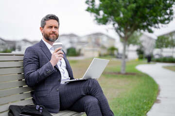 Business man sitting on bench in park, work on laptop, drink coffee. Businessman online work from laptop. Business man rest and reading news in american neighborhood. Business man is relax on bench.