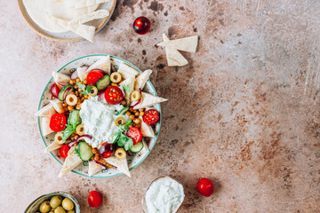 Mediterranean bowl with fried chickpeas, pita, olives, fresh vegetables and tzatziki sauce over beige rustic background