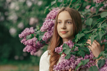 Young pretty dreaming girl standing among blooming branches of lilac in spring garden or park at sunset, warm cozy outdoor lifestyle portrait, idea of beauty, freshness, youth, relax and naturalness