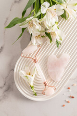 A rose quartz cosmetic roller massager, a gua sha scraper and astromeria flowers lie on a white...