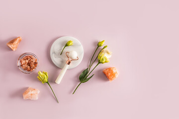 A professional rose quartz massager for facial care at home lies on a white marble round podium and sea salt in a bowl. Top view. austoma flowers