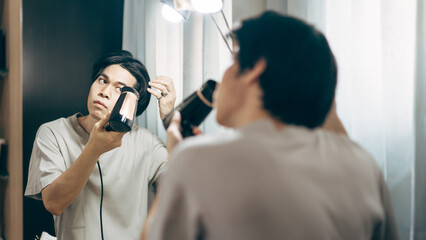 Young adult asian man using hair dryer blow for hairstyle in this mirror reflection at home