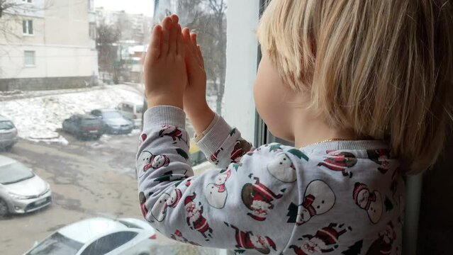 A little boy with blonde hair sits by the window on the windowsill and looks out the window. Winter. New Year's Eve. The kid is dressed in a New Year's light with snowmen. Baby boy with blond hair is