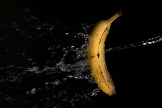 A banana against a dark background. Water splashes on the fruit from the side. The yellow of the banana is a contrast to the dark background.