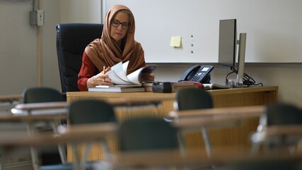 Happy smiling woman teacher wearing headscarf in empty school classroom working on assignments for students. - obrazy, fototapety, plakaty