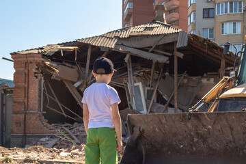 6 years old boy stands in front of a destroyed house. He looks at the destroyed building standing...