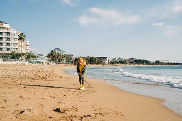 Obraz premium Boy in yellow rubber boots playing with stick and sand at the beach. School kid touching water at autumn winter sea. Child having fun with waves at the shore. Spring Holiday vacation concept.