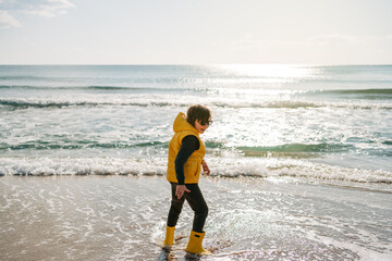 Boy in yellow rubber boots playing with sand at the beach. School kid touching water at autumn...