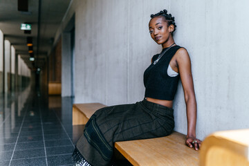 Portriat of african millennial woman posing indoor sitting on bench over gray wall background....
