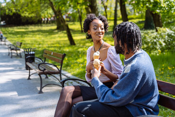 Happy black couple eating ice cream on a beach bench together outdoor at city park.