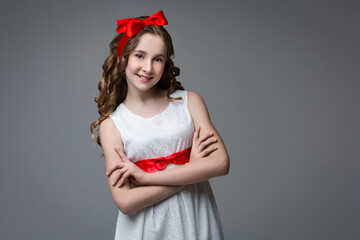 Beautiful teenage girl with long curly hair and red ribbon bow on head wearing white dress. Happy expression. Studio portrait on grey background. Copy space. - Powered by Adobe