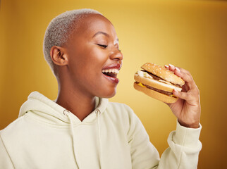 Burger, eating and young woman or student on studio, yellow background for restaurant, lunch and...