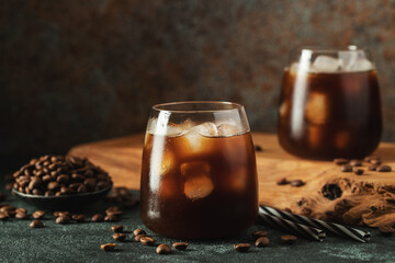 Ice coffee in a tall glass with cream poured over, ice cubes and beans on a dark concrete table.