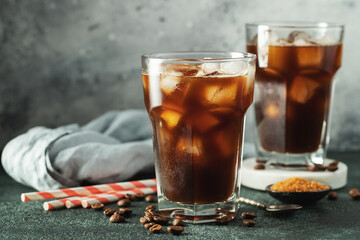 Ice coffee in a tall glass with cream poured over, ice cubes and beans on a dark concrete table.