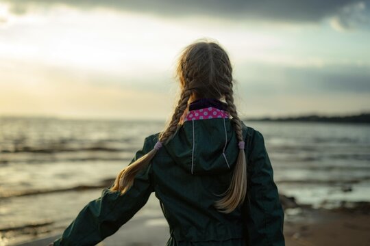 back view of caucasian blond girl with hairs styled with two plaits looking at sea on sunset