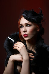 Beautiful young woman with smoky eyes and full red lips holding cigarette holder. Vintage head piece. Retro styling. Studio beauty shot. Copy space.