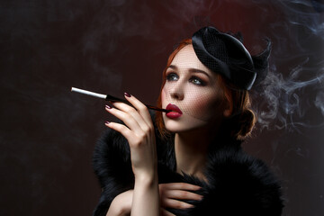 Beautiful young woman with smoky eyes and full red lips holding cigarette holder. Vintage head piece. Retro styling. Studio beauty shot over smoky background. Copy space.