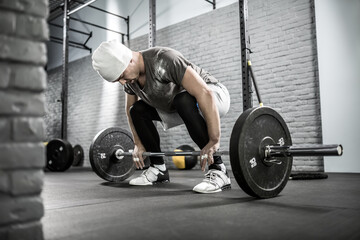 Fototapeta na wymiar Magnificent man with a beard starts to raise a barbell in the squat in the gym on the gray brick wall background. He wears sportswear, white sneakers and a white cap. Guy looks at the barbell.