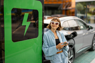 Young woman stands with a smart phone, waiting for her electric car to charge on a public charging...