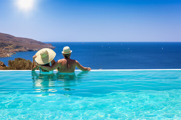 A loving couple with hats hugging at the edge of an infinity pool and enjoying the view to the blue, mediterranean sea during summer vacation time
