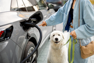 Woman pulling a charging plug out of her EV car, coming from a walk with her dog. Concept of...