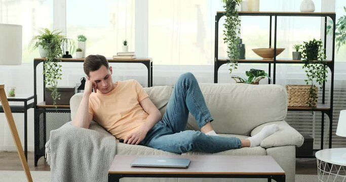 Caucasian man as he finds himself trapped in the doldrums of monotony at home. Seated on the sofa, his tired gaze and slouched posture reflect his longing for excitement and stimulation.