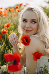 Obraz na płótnie Canvas Portrait of a blonde young woman in a field with wild red poppies, smiling, holding a red poppy.