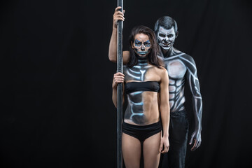 Grinning couple of pole dancers with horrific body-art stands next to a pylon on a dark background in the studio. They dressed in black sportswear and hold the right hands on the pylon. Horizontal.