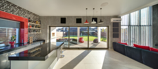 Fototapeta premium Interior in a modern style with a kitchen zone with a bar, concrete and brick walls, large windows and a door. On the right there is dark sofa with red pillows. Outside there is a terrace and a lawn.