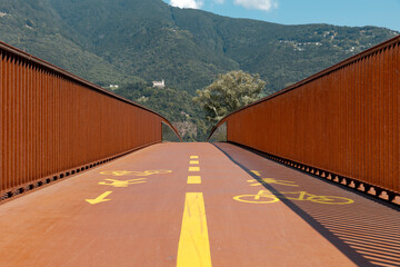 Front view of the pedestrian and bicycle path of a modern rusty bridge, with the Swiss Alps in Ticino in the background. Strong contrast between light and shadow, nice panorama